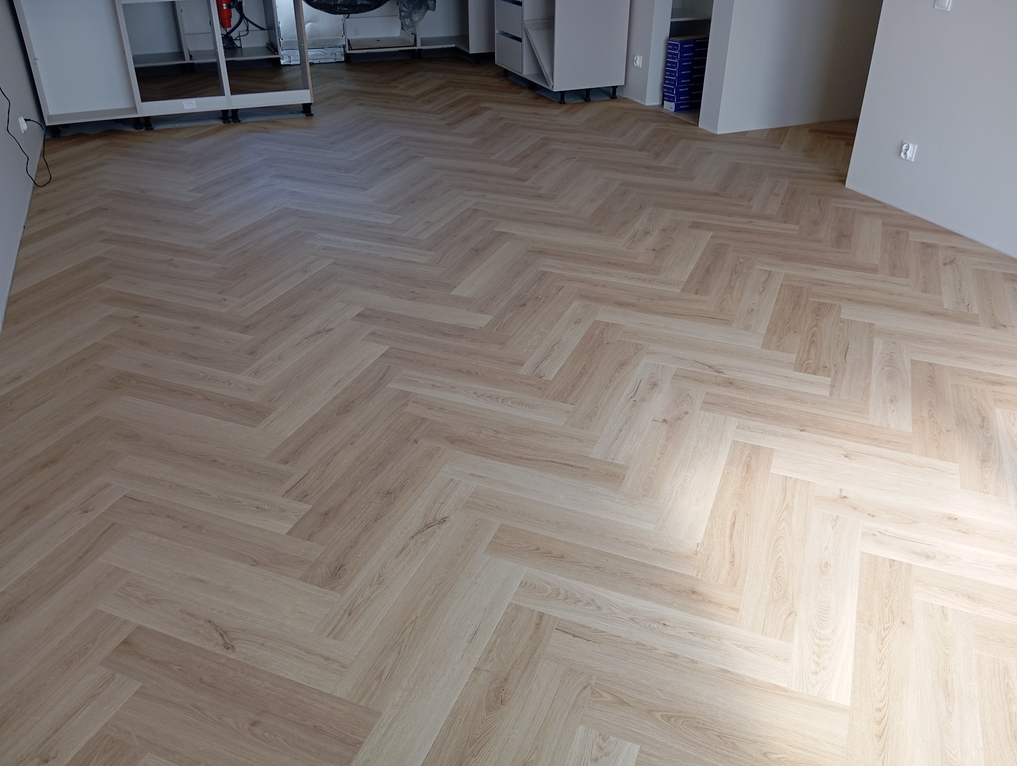 Jasna, drewnopodobna podłoga ułożona we wzór jodełki francuskiej, widoczne niedokończone szafki kuchenne w tle.