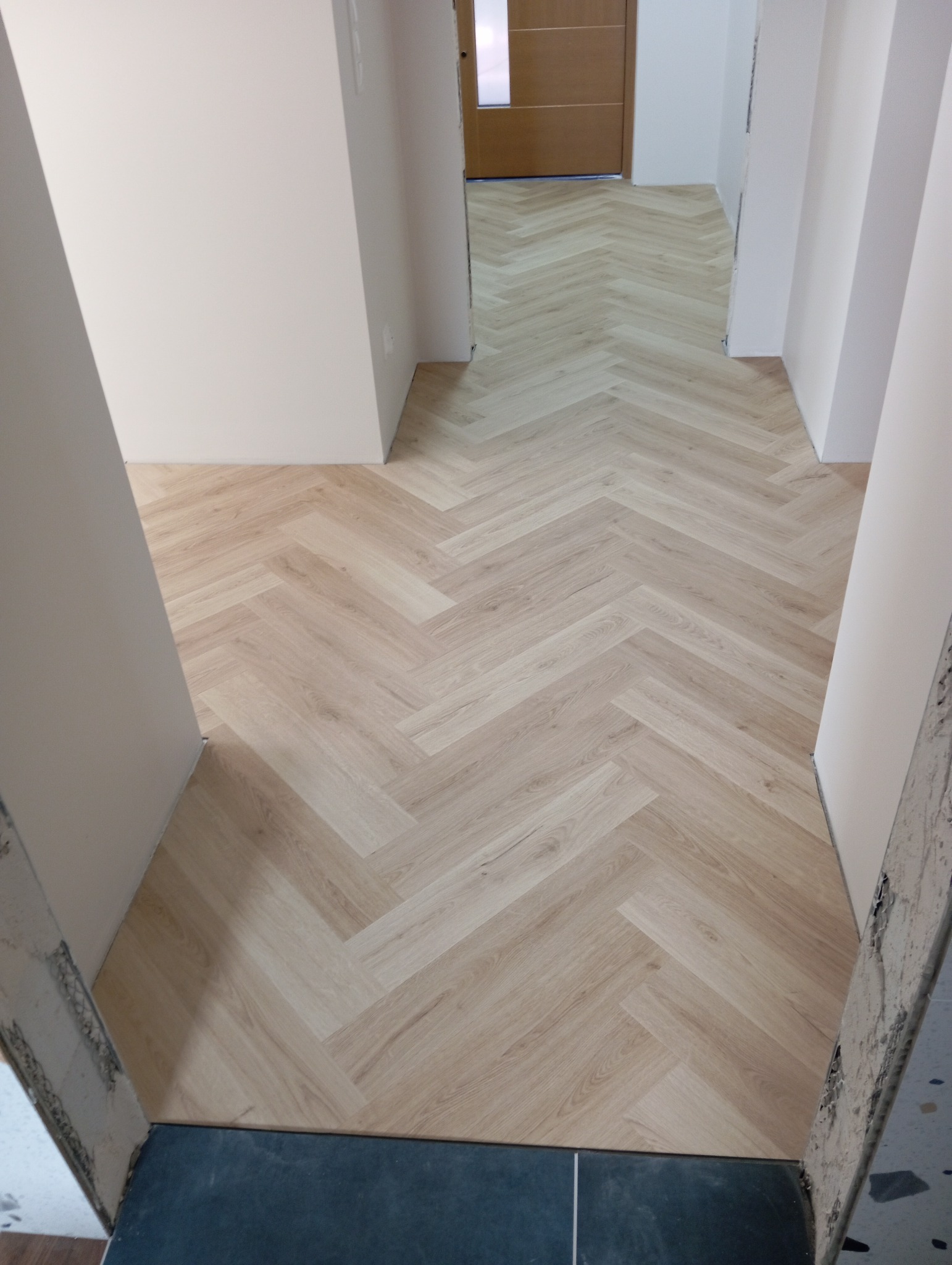 Jasne panele ułożone we wzór jodełki w korytarzu, widoczne niedokończone wykończenia ścian i drzwi w tle.