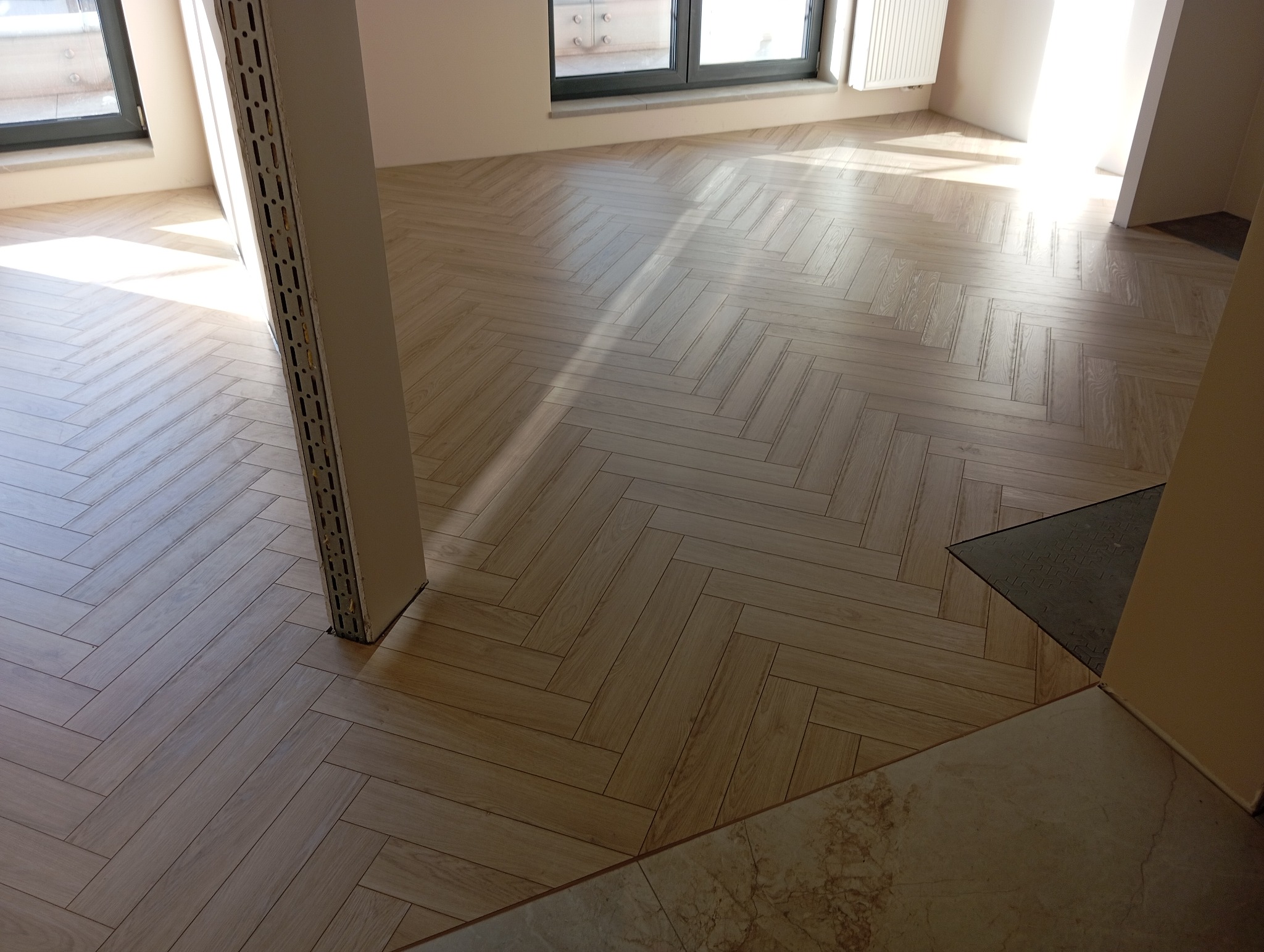 Jasne panele ułożone w jodełkę francuską w nowym wnętrzu, widoczny profil metalowy przy słupie, fragment marmurowej posadzki przy progu.