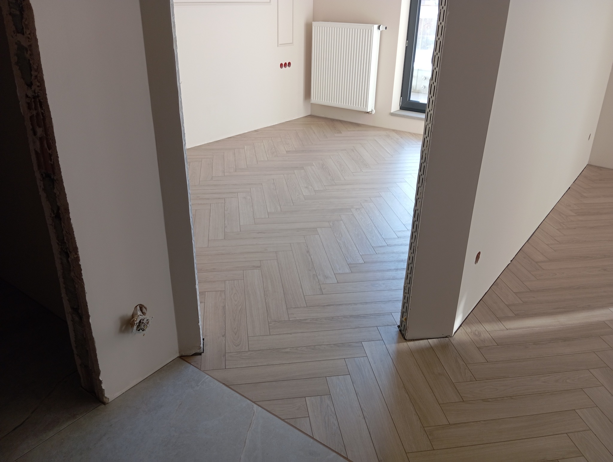 Ułożony parkiet w jodełkę w jasnym odcieniu drewna, widok z częściowo wykończonego pomieszczenia z widoczną instalacją elektryczną i grzejnikiem.