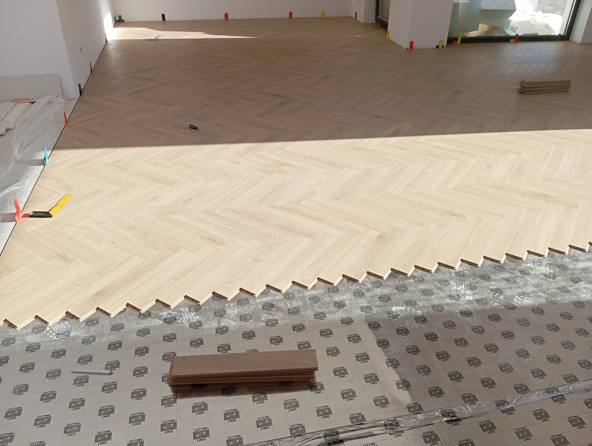 Montaż jasnego parkietu w jodełkę francuską, widoczna warstwa podkładowa z nadrukiem siatki, narzędzie w tle, niedokończony fragment podłogi.