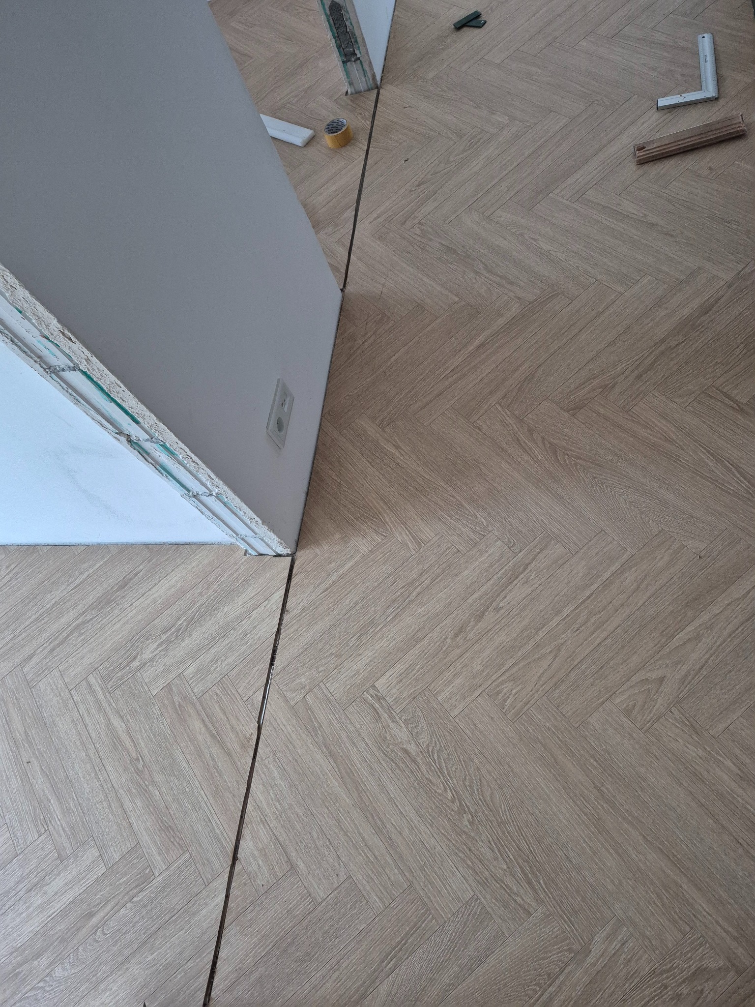 Jasne panele ułożone we wzór jodełki, widoczne narzędzia i fragment ściany z gniazdkiem. Układanie parkietu w trakcie, widok z góry.