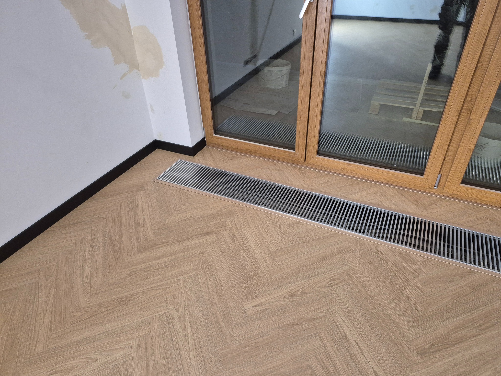 Ułożona podłoga z paneli w jodełkę, z widoczną metalową kratką wentylacyjną przy oknie balkonowym. Ściana z widocznymi śladami po szpachlowaniu.