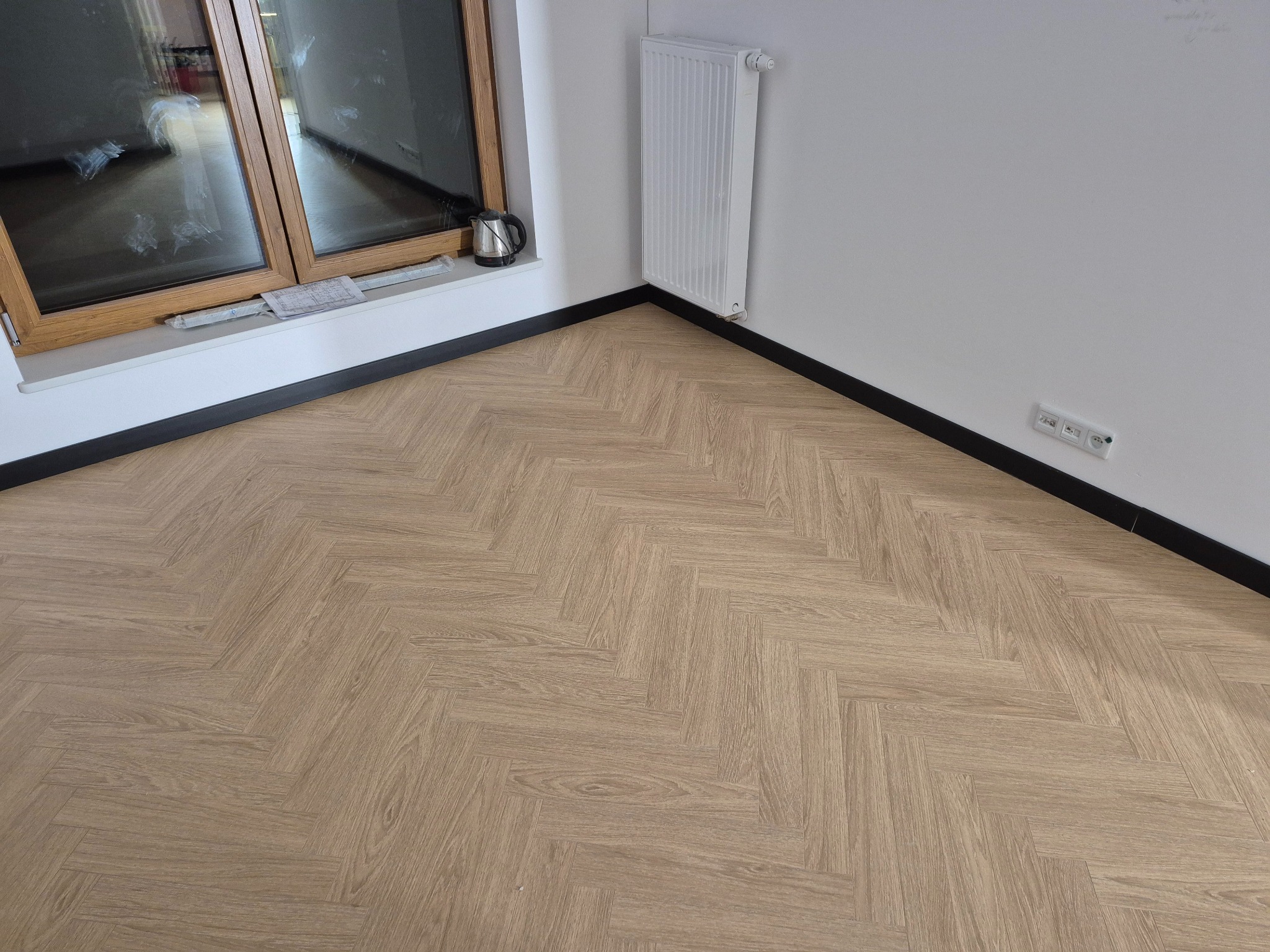 Jasna podłoga ułożona we wzór jodełki francuskiej w rogu pokoju z oknem, grzejnikiem i listwami przypodłogowymi. Widoczne gniazdka elektryczne na ścianie.