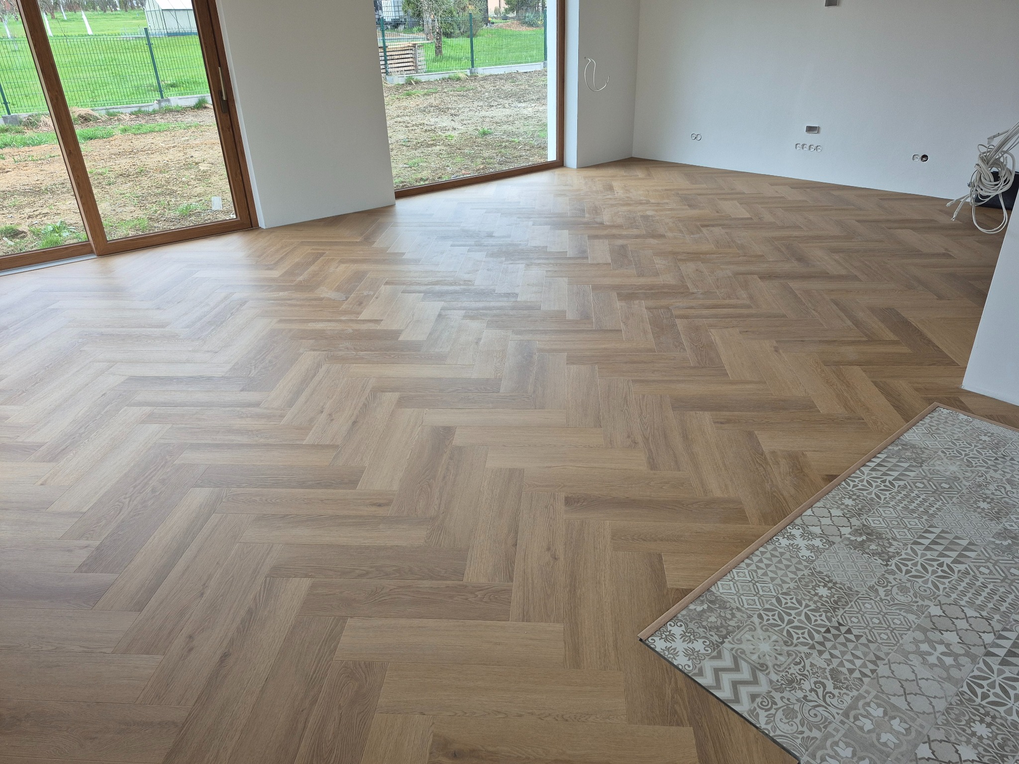 Ukończona podłoga z paneli ułożonych w jodełkę francuską w jasnym, pustym pokoju z dużymi oknami i fragmentem dywanu w rogu. Widoczne gniazdka elektryczne.