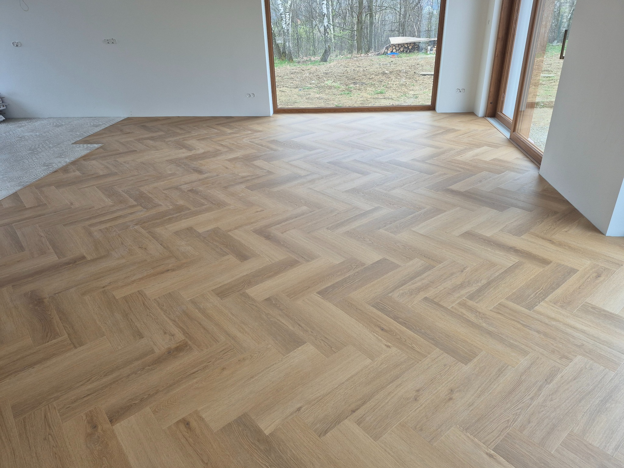 Jasne panele ułożone w jodełkę francuską, częściowo pokrywające podłogę w nowym wnętrzu z dużym oknem na ogród. Widoczny fragment nieukończonej podłogi.