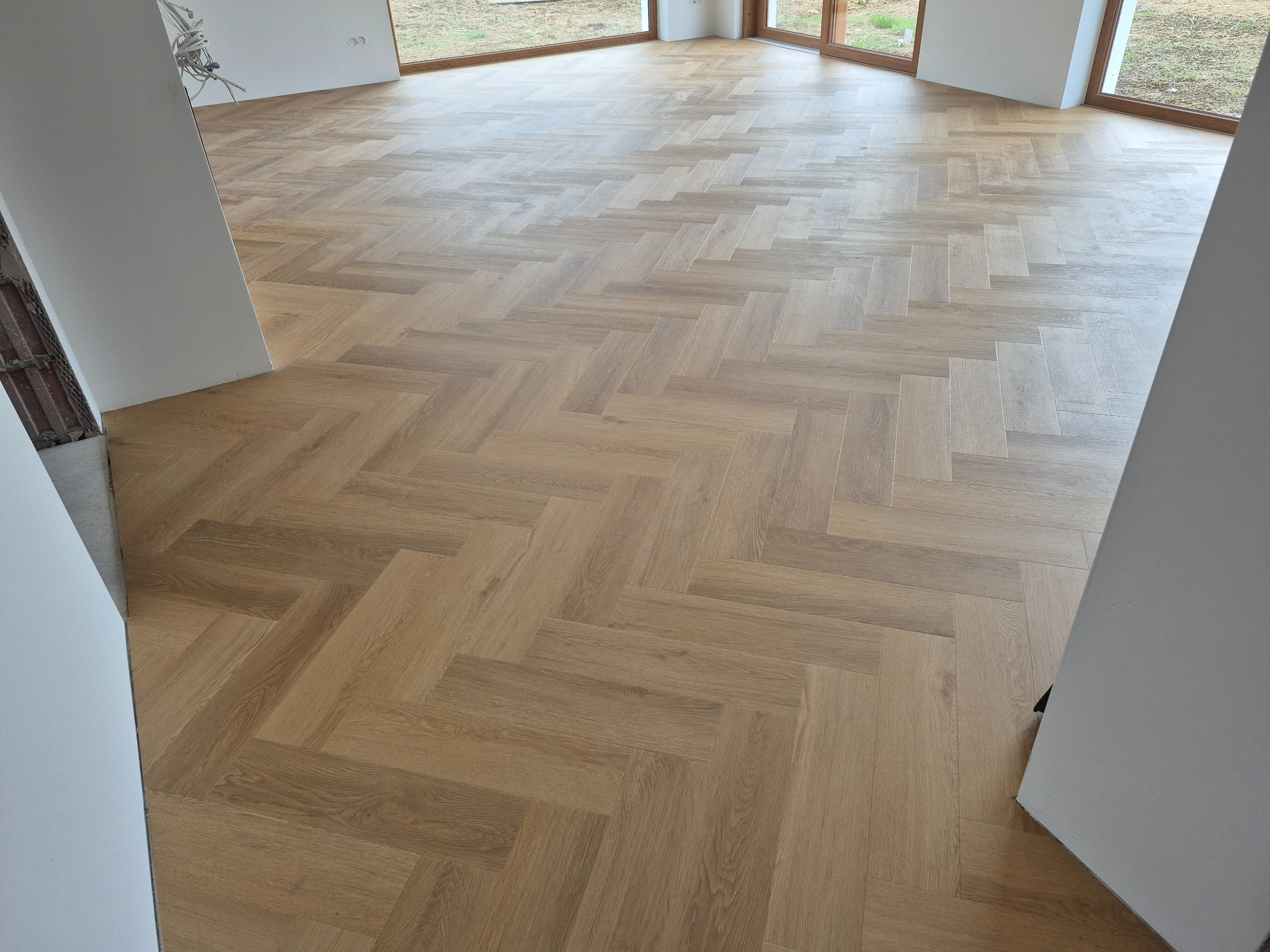 Jasna, drewniana podłoga ułożona w jodełkę francuską w nowym wnętrzu z dużym oknem i widocznymi przewodami elektrycznymi przy ścianie.
