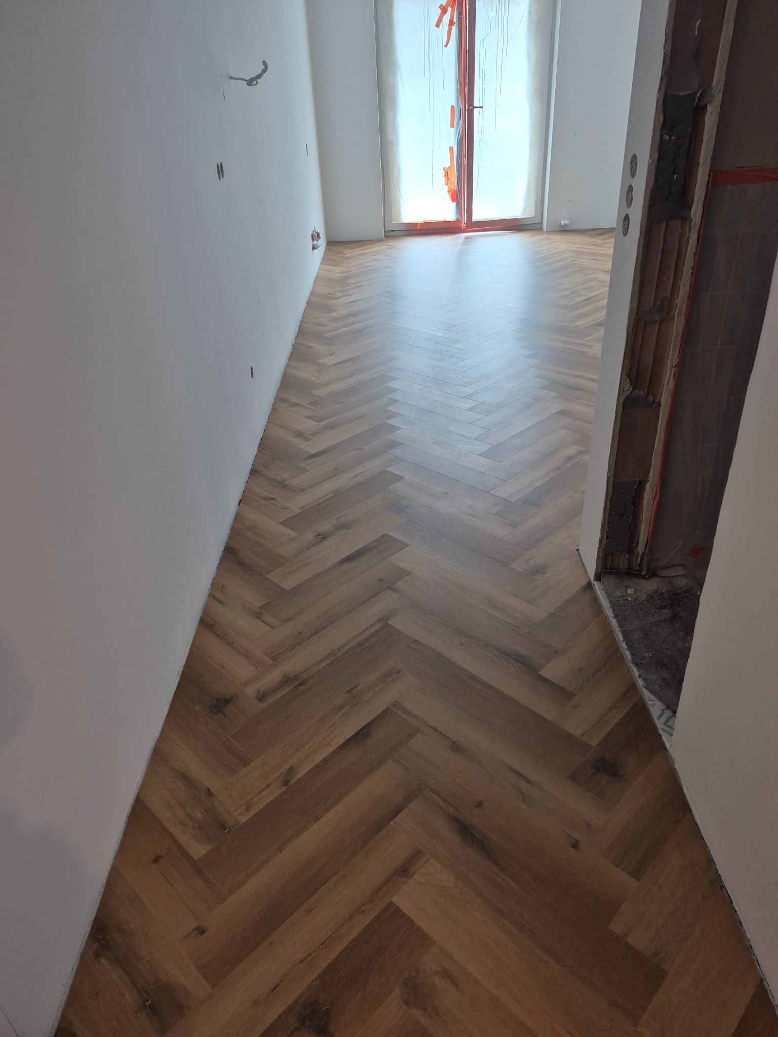 Ułożona jodełka z paneli w jasnym korytarzu, widok w perspektywie, surowe ściany, w tle okno balkonowe z folią zabezpieczającą.