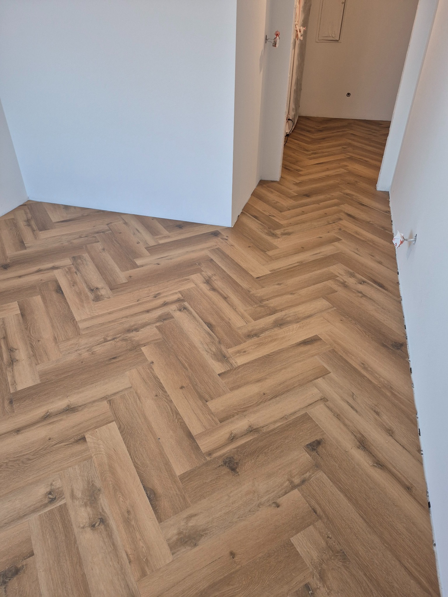 Jasne panele ułożone w jodełkę francuską, widok z góry. Podłoga w odcieniach brązu, świeżo położona, przy białych ścianach i przejściu do kolejnego pomieszczenia.