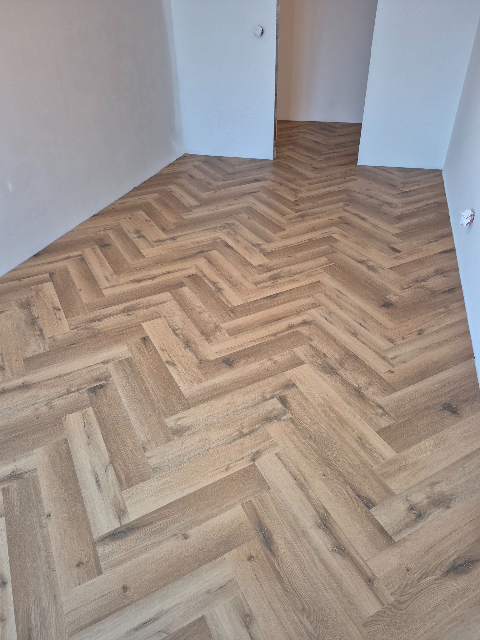 Jasne panele ułożone w jodełkę francuską, widok z góry na fragment pomieszczenia z białymi ścianami. Nowoczesne wykończenie podłogi.