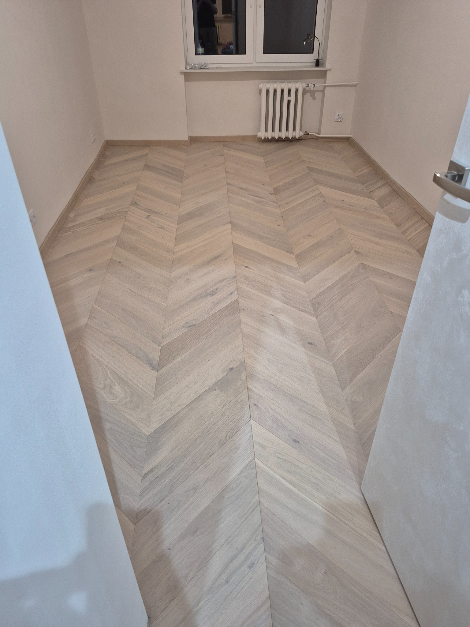 Jasny parkiet ułożony we wzór jodełki francuskiej wypełnia pokój z grzejnikiem i oknem. Widok zza uchylonych drzwi na podłogę po renowacji.