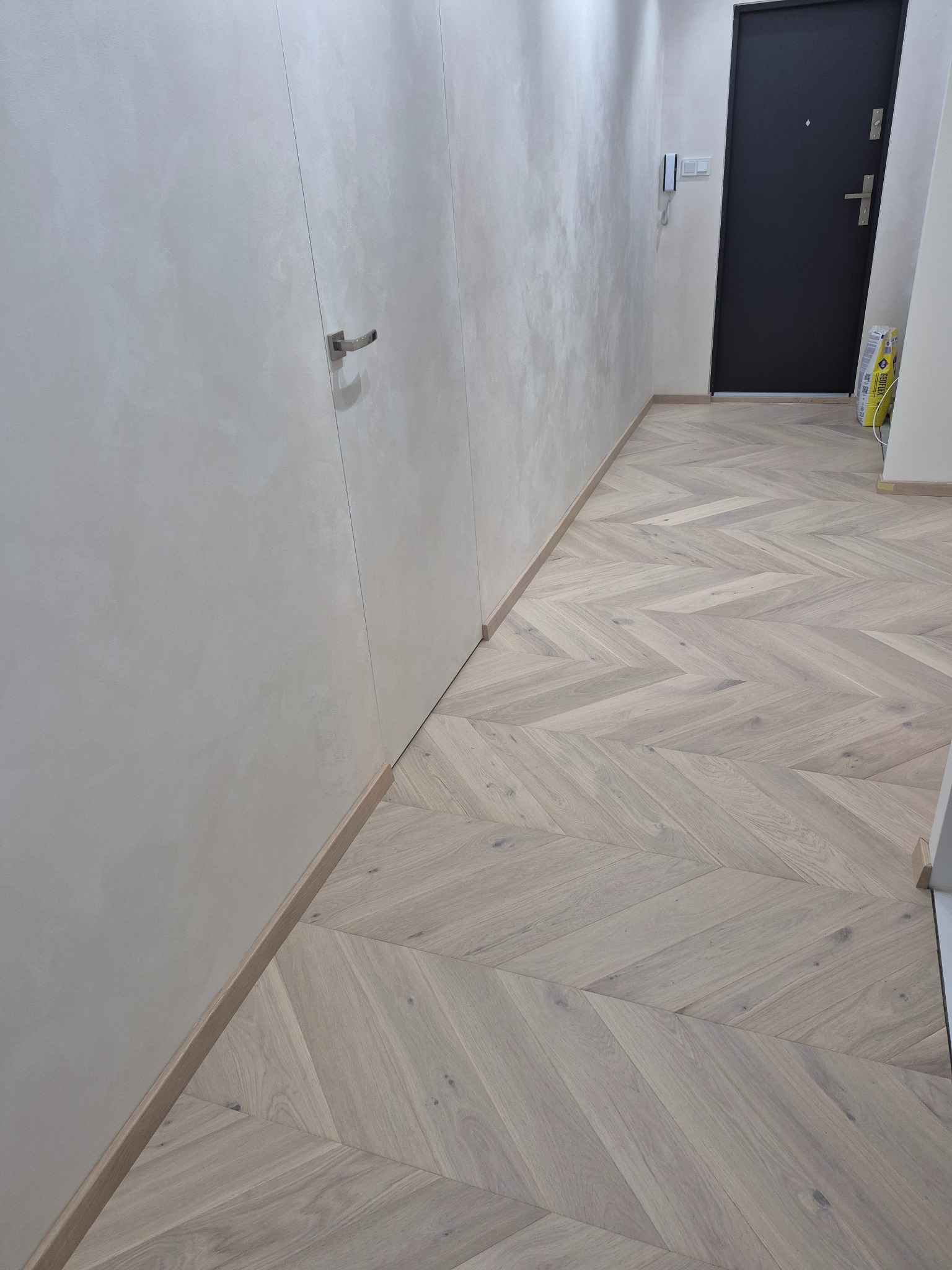 Jasny parkiet ułożony we wzór jodełki francuskiej w korytarzu z szarą ścianą i czarnymi drzwiami wejściowymi. Widoczne listwy przypodłogowe i fragment ściany.