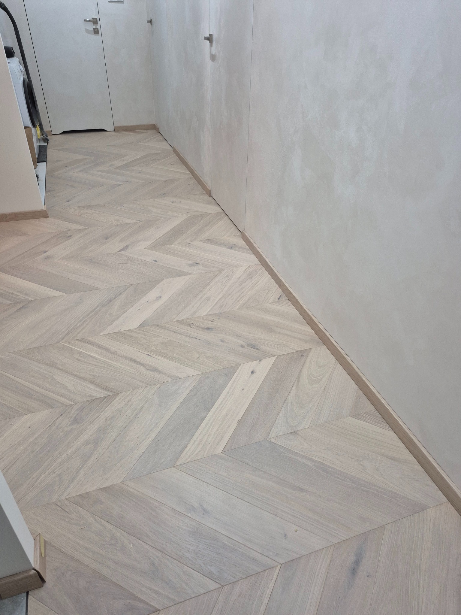 Jasna, drewniana podłoga ułożona we wzór jodełki francuskiej w korytarzu. Minimalistyczne wnętrze z białymi drzwiami i ścianami o fakturze betonu.