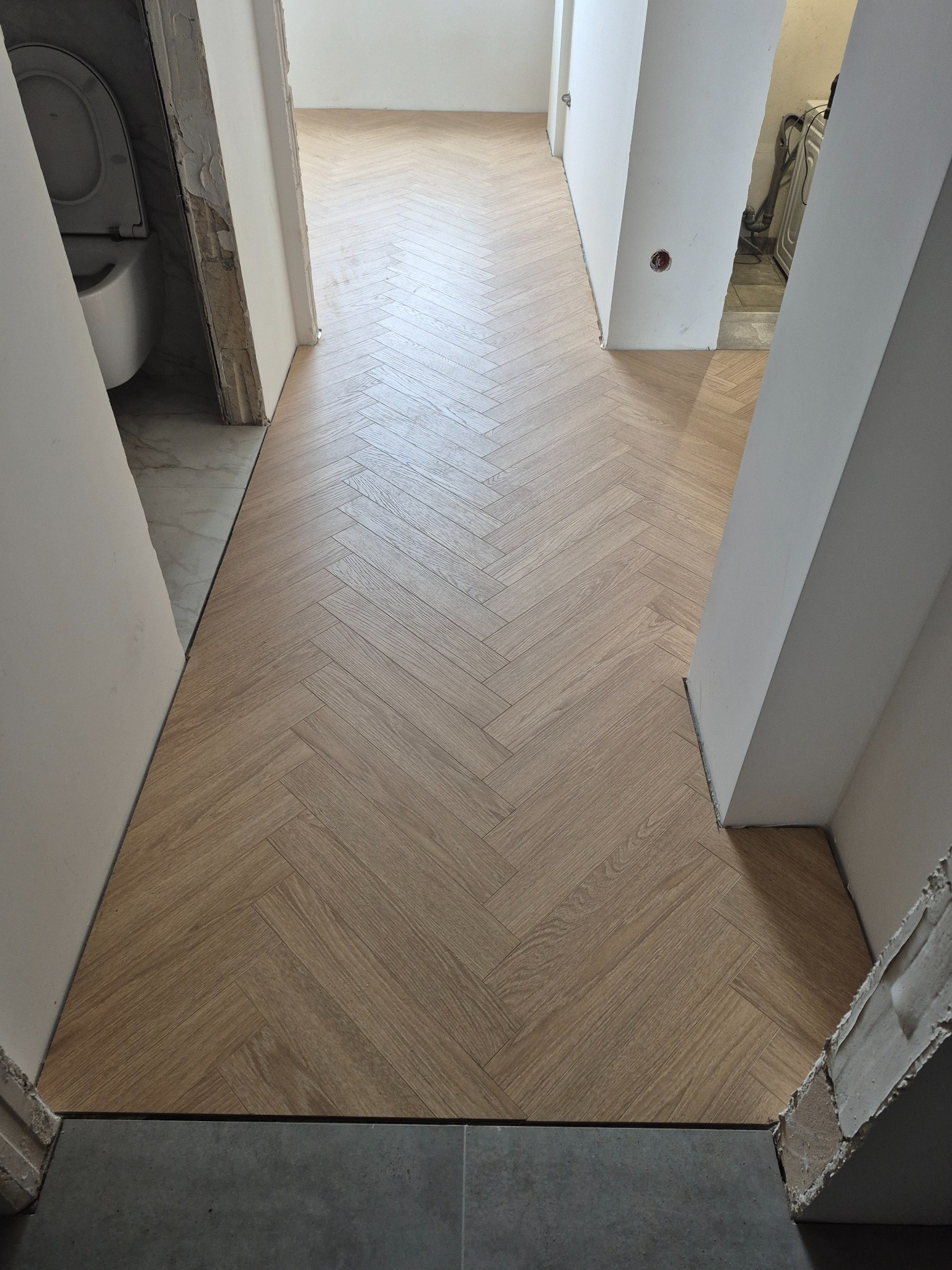 Jasne panele ułożone w jodełkę w przejściu między pokojami, widoczne niedokończone wykończenia ścian i podłogi w sąsiednich pomieszczeniach.