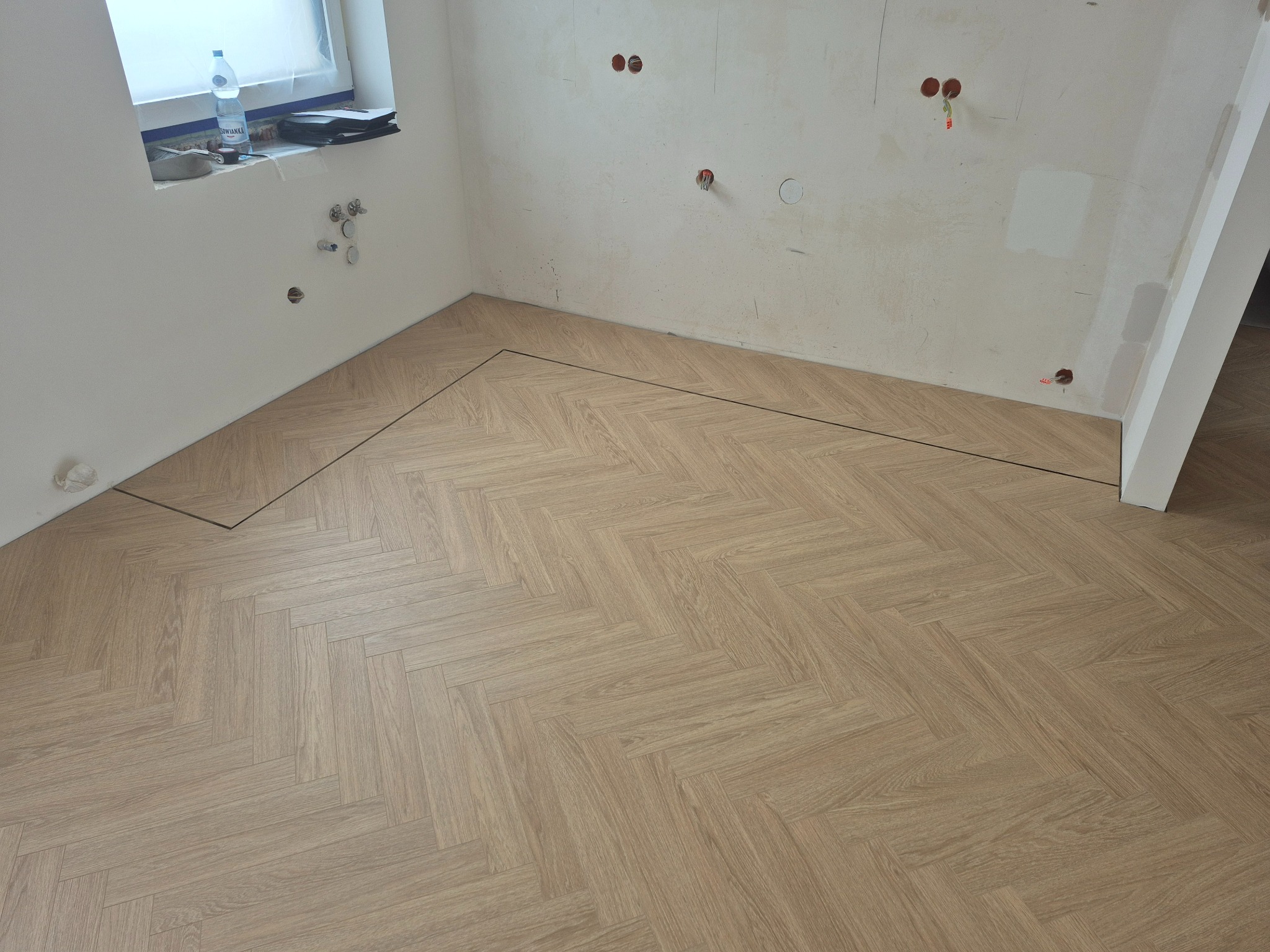 Jasny parkiet ułożony we wzór jodełki w pokoju z białymi ścianami. Widoczne przygotowania do dalszych prac wykończeniowych. Butelka wody na parapecie.