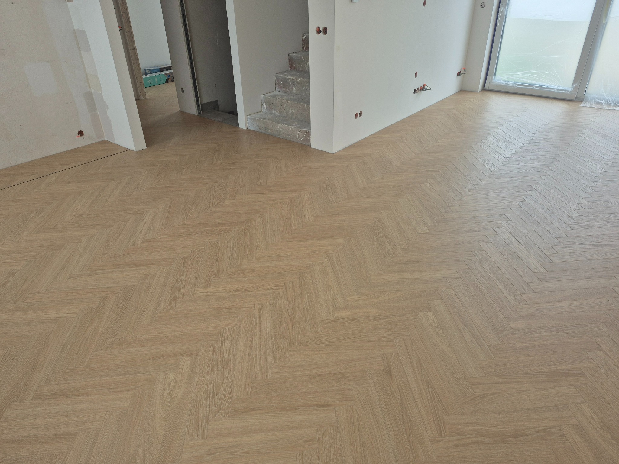 Jasnobrązowa podłoga ułożona w jodełkę w surowym wnętrzu z widocznymi schodami i oknem zabezpieczonym folią. Podłoże świeżo po montażu.