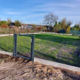 Nowo zainstalowane, grafitowe ogrodzenie panelowe z betonową podmurówką i furtką na tle zielonego trawnika i błękitnego nieba. Widoczne ślady prac ziemnych przy podmurówce.