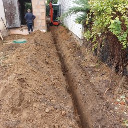 Prace Ziemne Adam Mroziński - Wąski wykop w ziemi wzdłuż ściany budynku, z małą koparką w tle i dwoma osobami w otwartych drzwiach piwnicy.