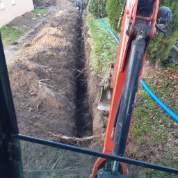 Prace Ziemne Adam Mroziński - Wąski wykop w ziemi wykonany koparką, widok z kabiny operatora, w tle pracujący robotnik.