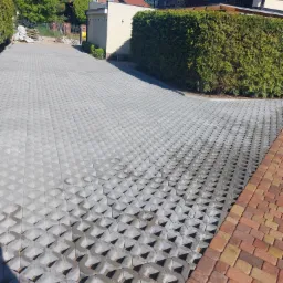 Ułożona szara ażurowa kostka brukowa na podjeździe, częściowo wypełniona piaskiem, z widocznym fragmentem czerwono-żółtej kostki po prawej stronie, w tle dom z balkonem i zielony żywopłot.