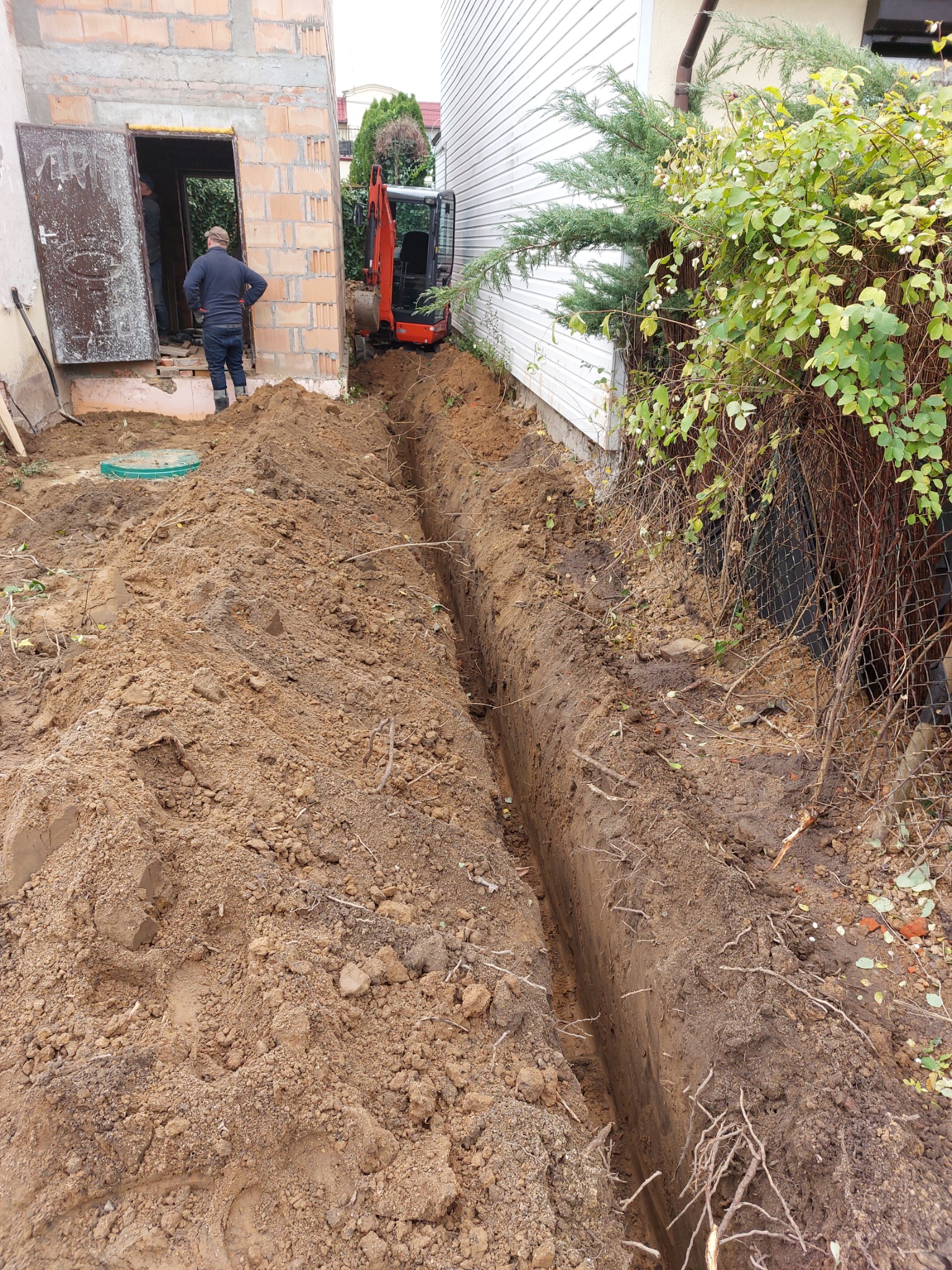 Wąski wykop w ziemi wzdłuż ściany budynku, z małą koparką w tle i dwoma osobami w otwartych drzwiach piwnicy.