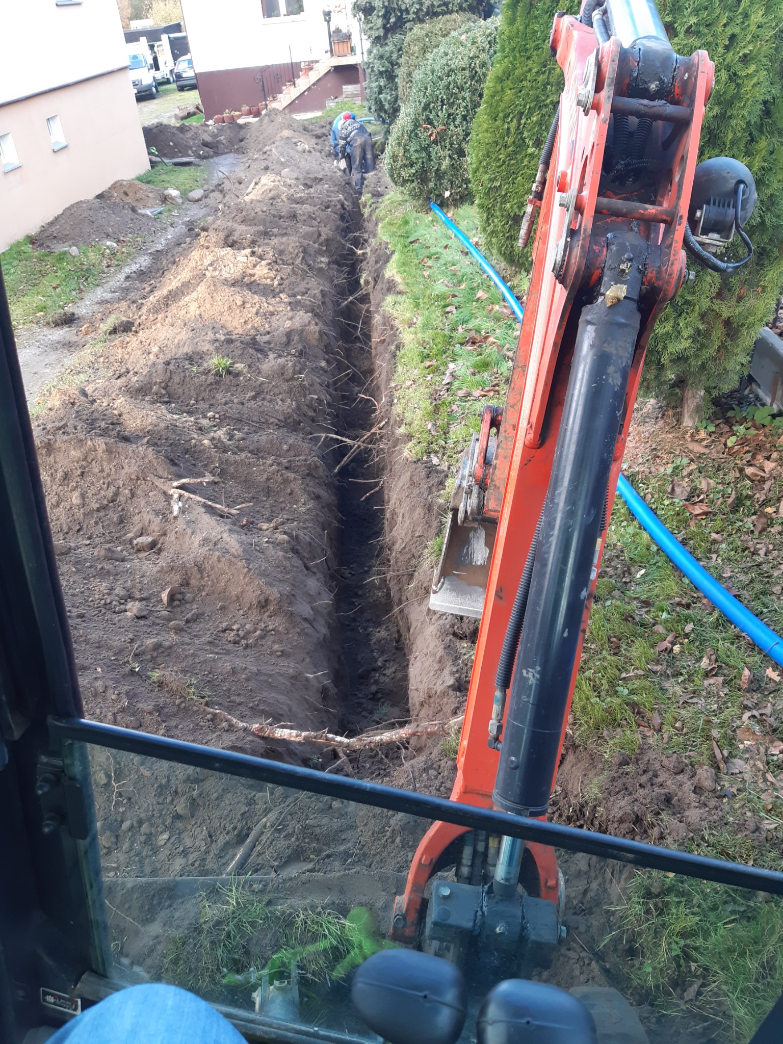 Wąski wykop w ziemi wykonany koparką, widok z kabiny operatora, w tle pracujący robotnik.