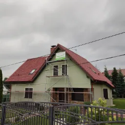 Dom jednorodzinny w trakcie malowania elewacji na jasnozielony kolor, widoczne rusztowania i częściowo wykończona elewacja z elementami kamienia dekoracyjnego, czerwony dach i szara metalowa brama.