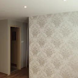 Wykończone wnętrze pokoju z wzorzystą tapetą w odcieniach beżu i brązu na jednej ścianie, gładką szarą ścianą obok drewnianych drzwi i parkietem na podłodze.