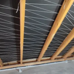 Wnętrze poddasza z widoczną konstrukcją drewnianą dachu, czarną membraną dachową i białymi sznurkami rozciągniętymi pomiędzy krokwiami, przygotowanymi do montażu płyt kartonowo-gipsowych.