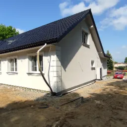 Świeżo otynkowany dom jednorodzinny z czarną dachówką, widoczna elewacja w jasnym odcieniu szarości, białe ramy okienne, element dekoracyjny narożnika, grunt wokół domu w trakcie prac ziemnych,...