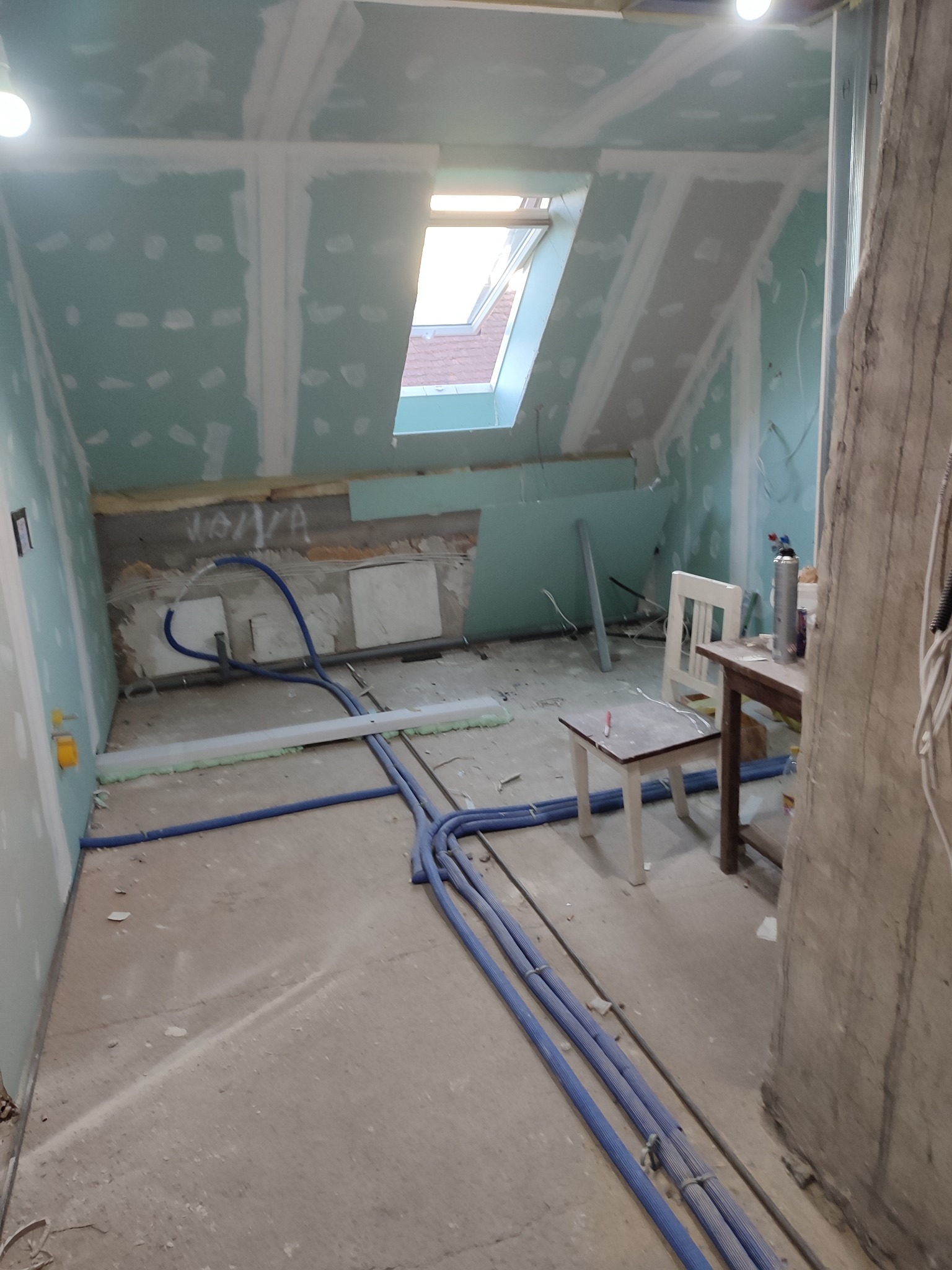 Poddasze w trakcie remontu: ściany i sufit pokryte płytami kartonowo-gipsowymi, widoczne niebieskie rury instalacji, okno dachowe wpuszczające światło.