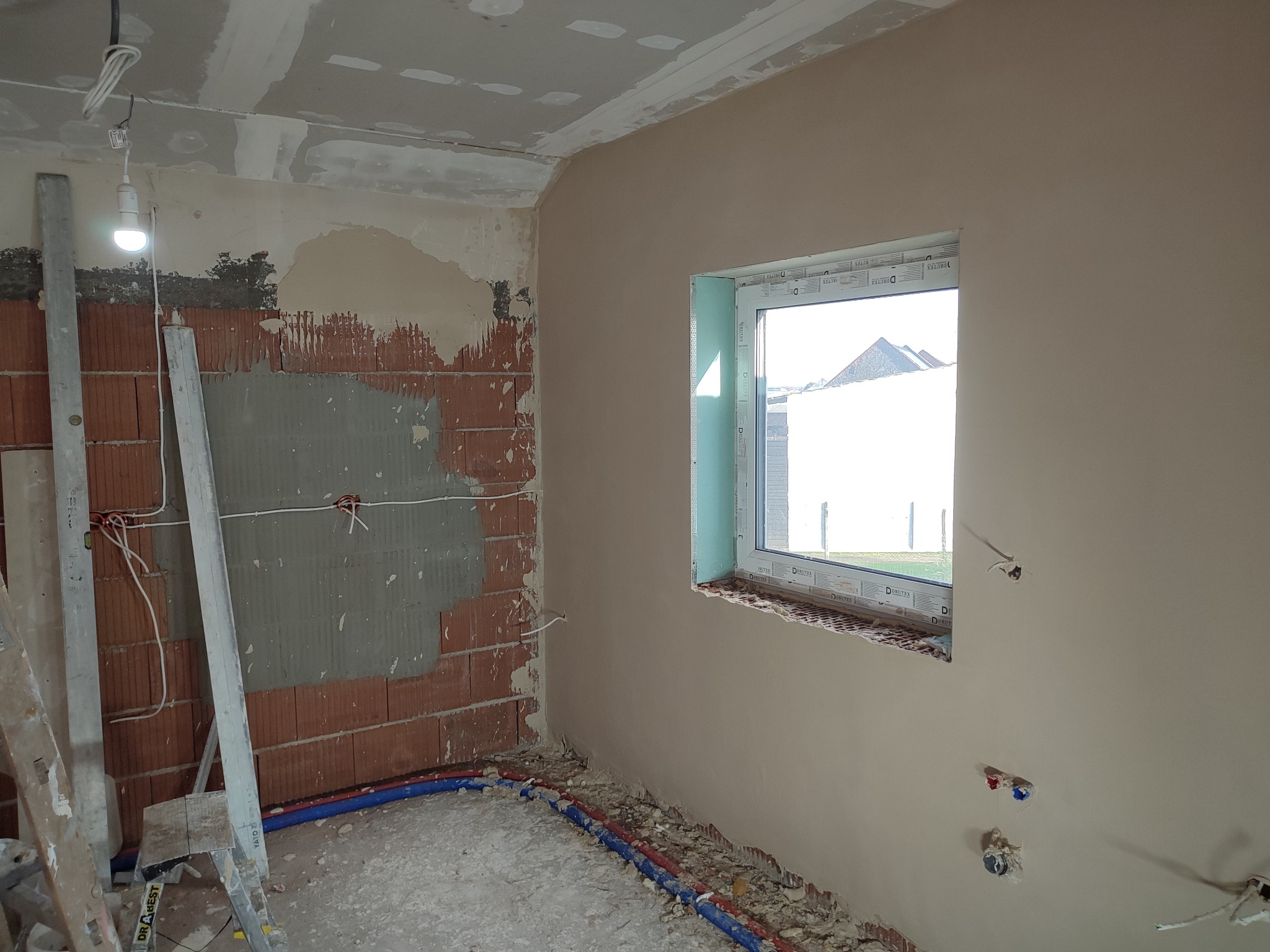 Wnętrze w trakcie prac wykończeniowych: częściowo otynkowana ściana z oknem, odkryta cegła, instalacje elektryczne i hydrauliczne, drabina oparta o ścianę.