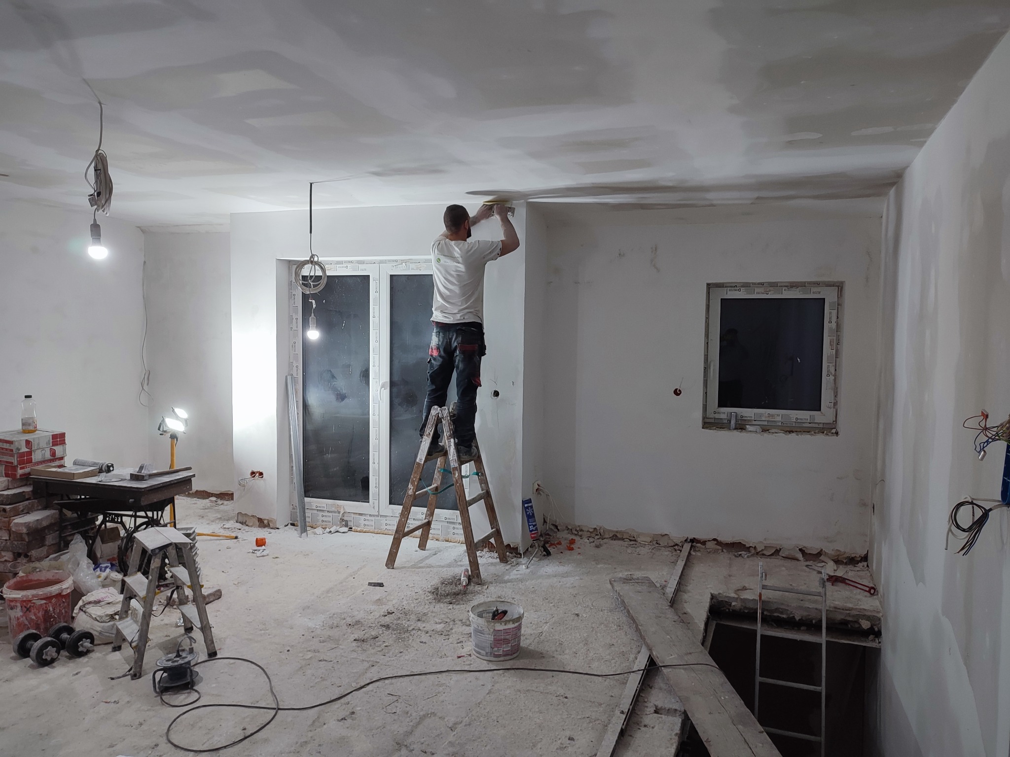 Mężczyzna na drabinie szlifuje sufit w trakcie remontu mieszkania, widoczne narzędzia, przewody elektryczne i okno balkonowe.