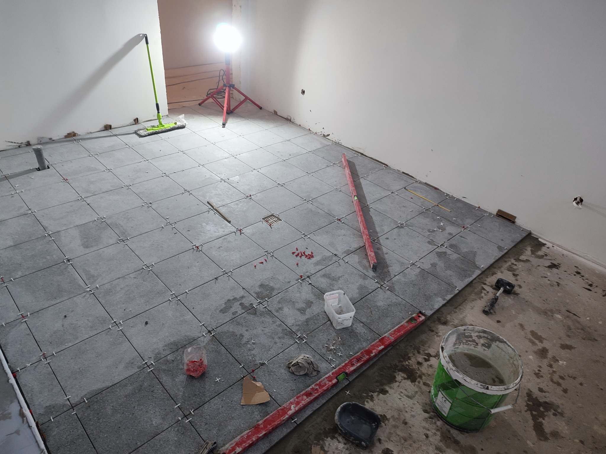 Układanie szarych płytek podłogowych w trakcie remontu, widoczne poziomice, młotek, wiadro z zaprawą, krzyżyki dystansowe i lampa budowlana na trójnogu.