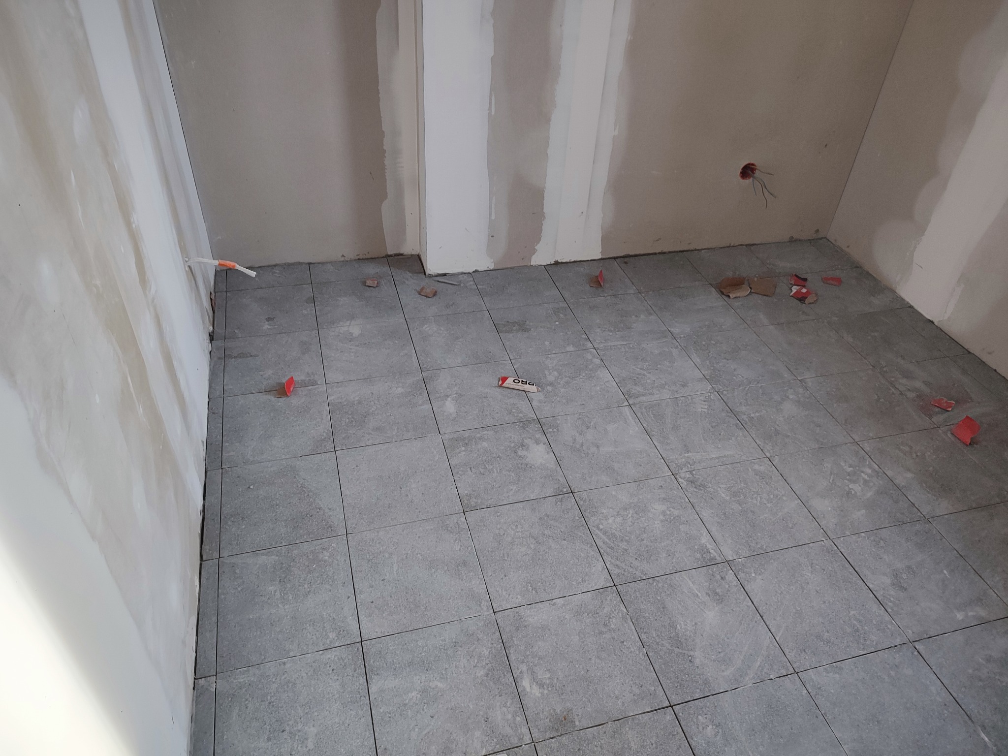 Szare płytki podłogowe ułożone w pomieszczeniu w trakcie remontu, widoczne fragmenty płyt karton-gips na ścianach i resztki po cięciu płytek na podłodze.