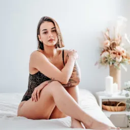 Kobieta w czarnym koronkowym body siedzi na łóżku, obejmując kolana, z tatuażami na ramionach i krótkimi brązowymi włosami, w tle wazon z kwiatami i świece.