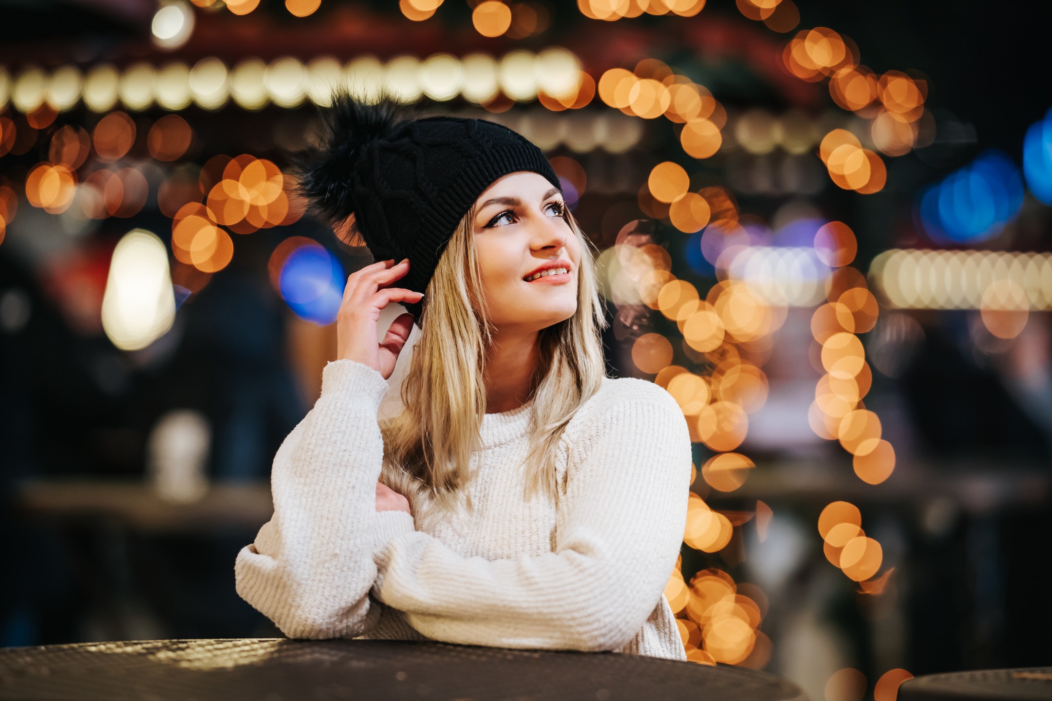 Portret uśmiechniętej kobiety w czapce z pomponem, opierającej się o stół na tle rozmytych światełek.