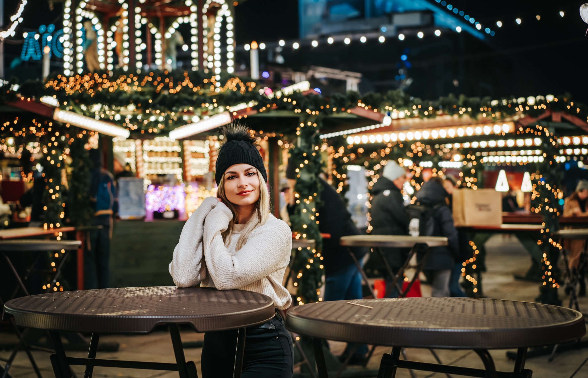 Kobieta w czapce z pomponem pozuje na tle świątecznego jarmarku z oświetlonymi straganami, opierając się o brązowy stolik.
