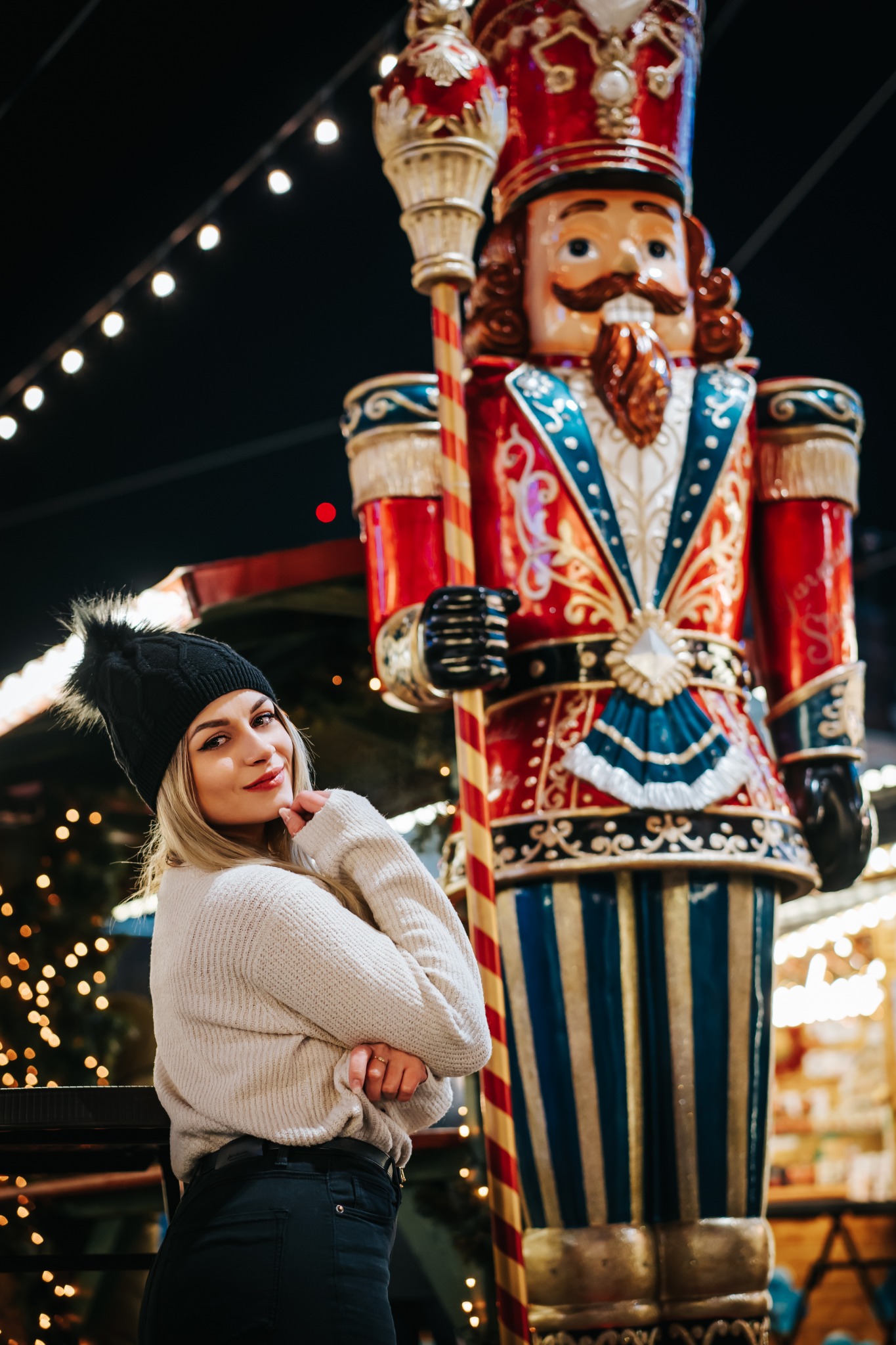 Uśmiechnięta blondynka w czapce z pomponem pozuje obok dużego, dekoracyjnego dziadka do orzechów na tle światełek.