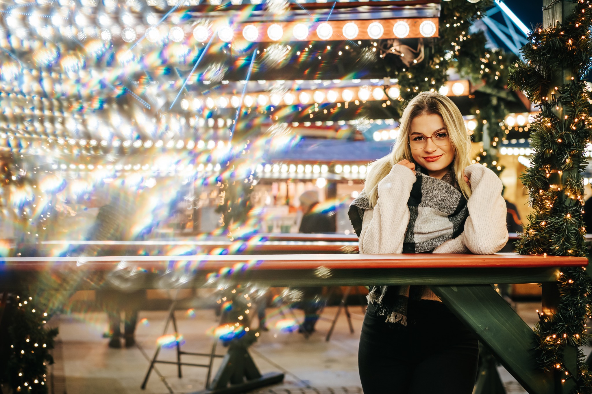 Uśmiechnięta blondynka w okularach, oparta o zielony stół z girlandą świąteczną, w tle rozświetlona dekoracja jarmarku bożonarodzeniowego.
