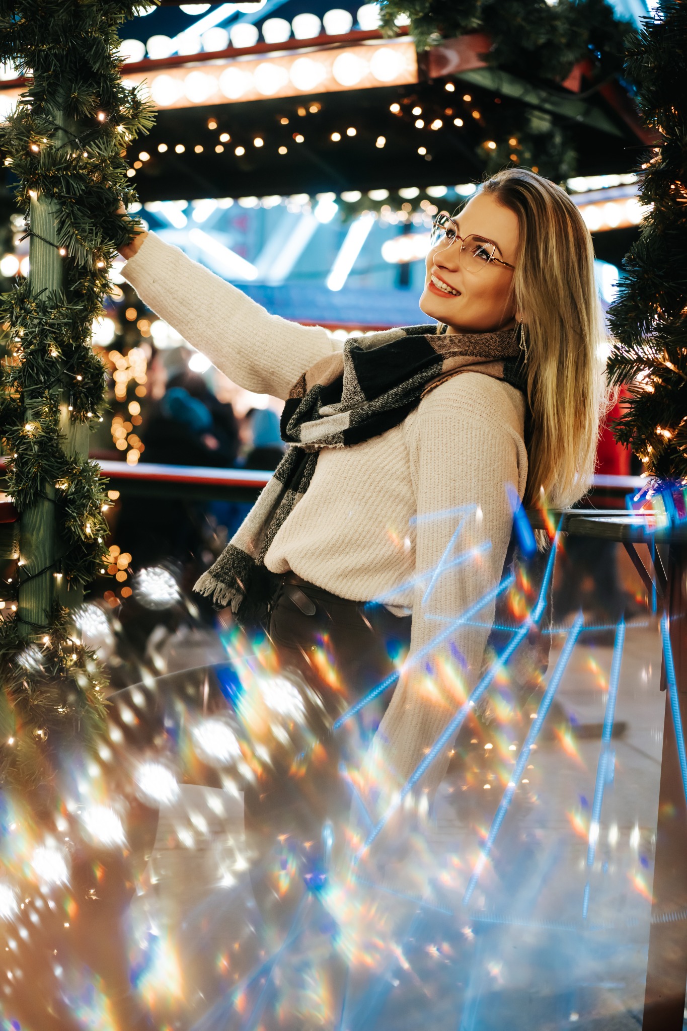 Uśmiechnięta blondynka w okularach, swetrze i szalu pozuje przy świątecznej dekoracji z lampkami i choinkowymi gałązkami, widoczne efekty świetlne.