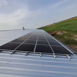 Panele słoneczne zamontowane na metalowym dachu budynku gospodarczego, z widokiem na zielone pola w tle.