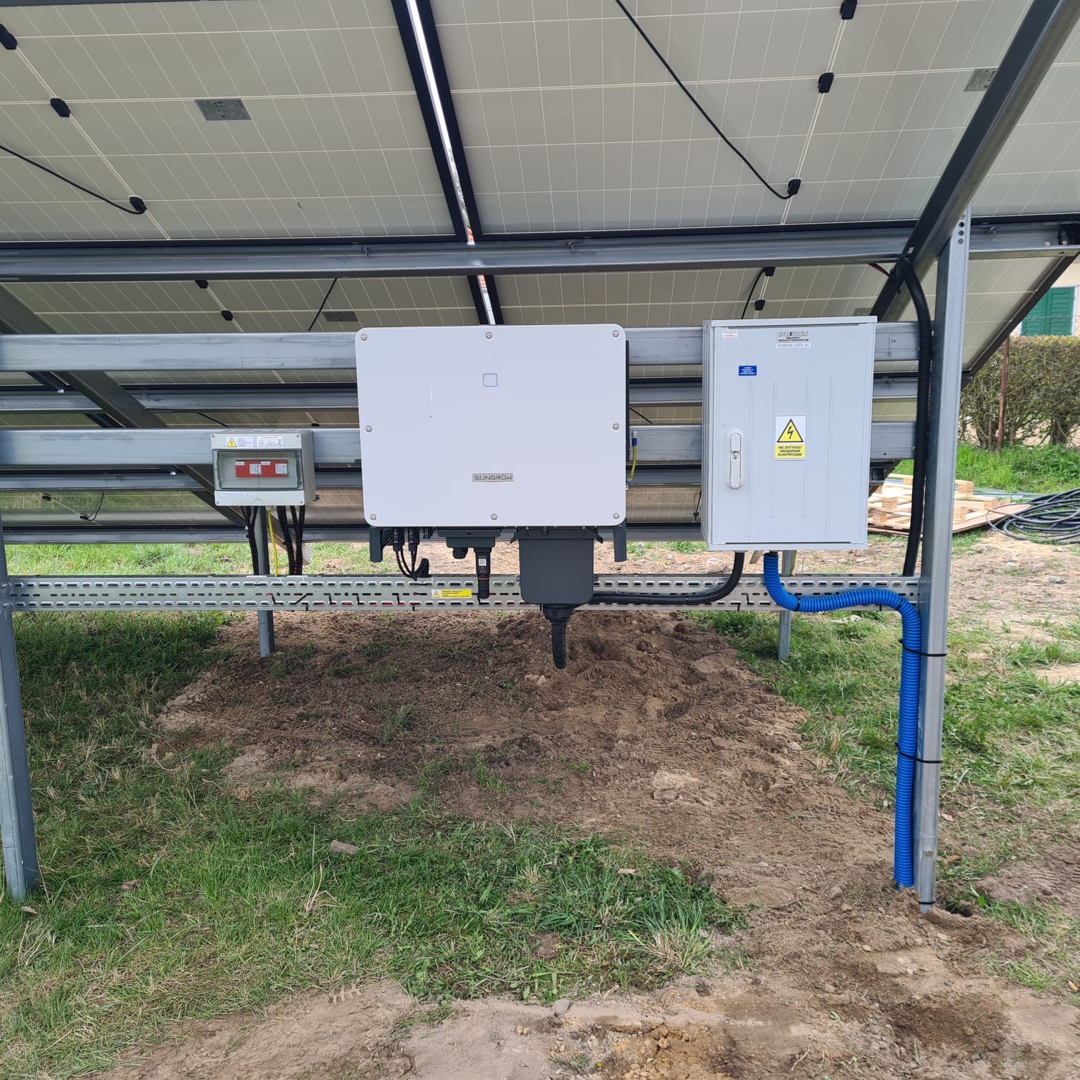Instalacja paneli słonecznych z widocznymi inwerterami i skrzynką zabezpieczeń na metalowej konstrukcji, z fragmentem trawnika w tle.