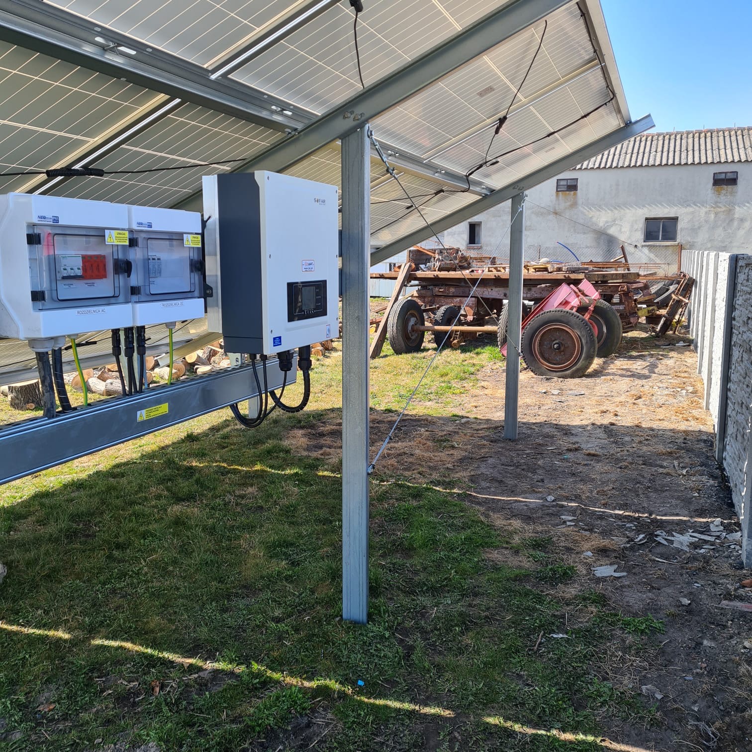 Instalacja paneli słonecznych z falownikiem i rozdzielnicą AC na trawiastym terenie, w tle budynek gospodarczy i sprzęt rolniczy.