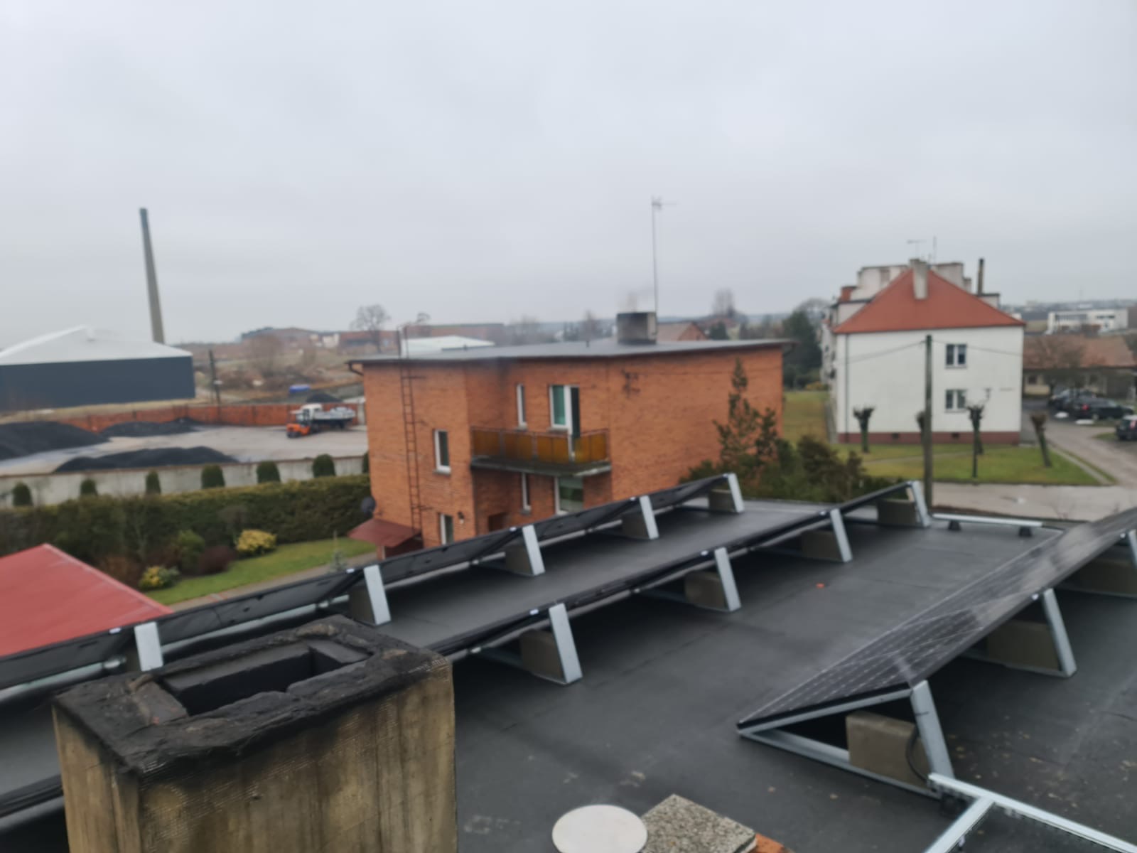 Widok z dachu w Pleszewie na instalację paneli słonecznych, z kominem na pierwszym planie i budynkami w tle, pochmurny dzień.
