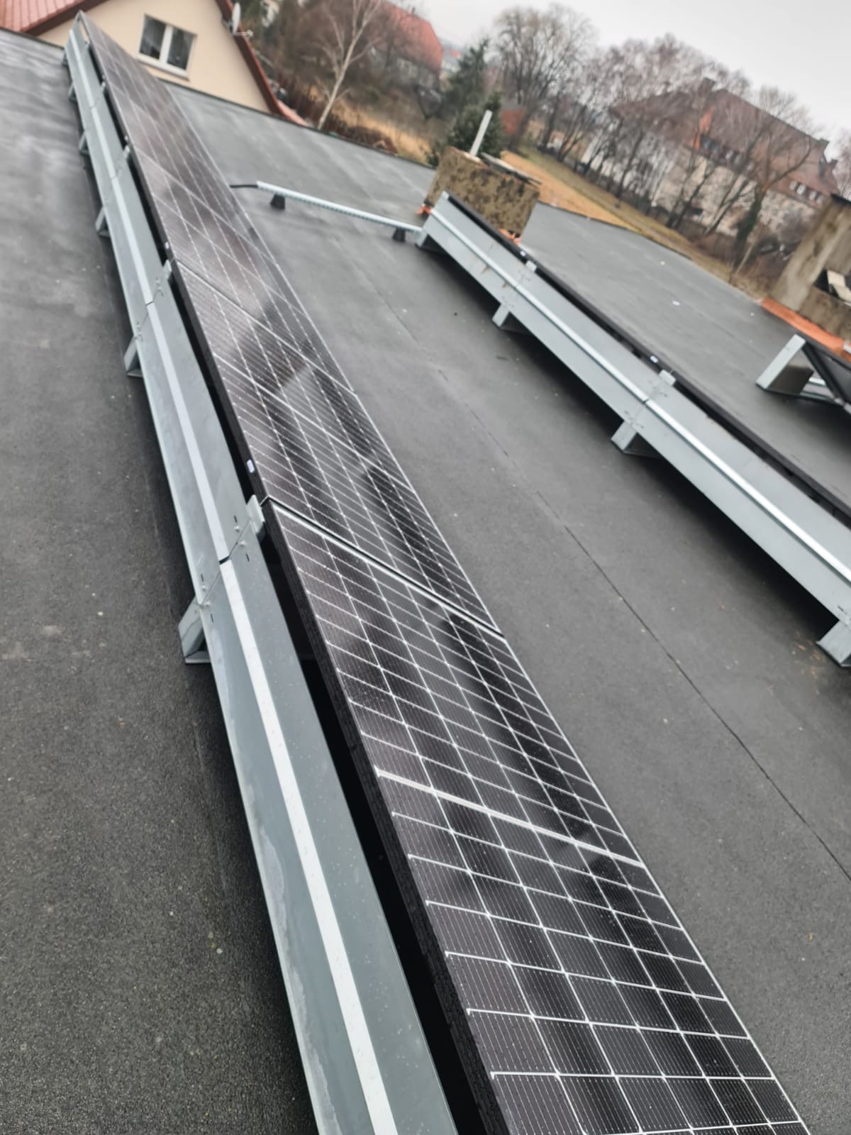 Rząd paneli fotowoltaicznych na płaskim dachu z widocznymi elementami montażowymi i budynkami w tle, wykonany w pochmurny dzień.