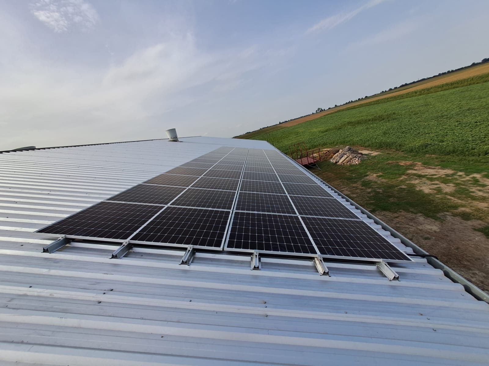 Panele słoneczne zamontowane na metalowym dachu budynku gospodarczego, z widokiem na zielone pola w tle.