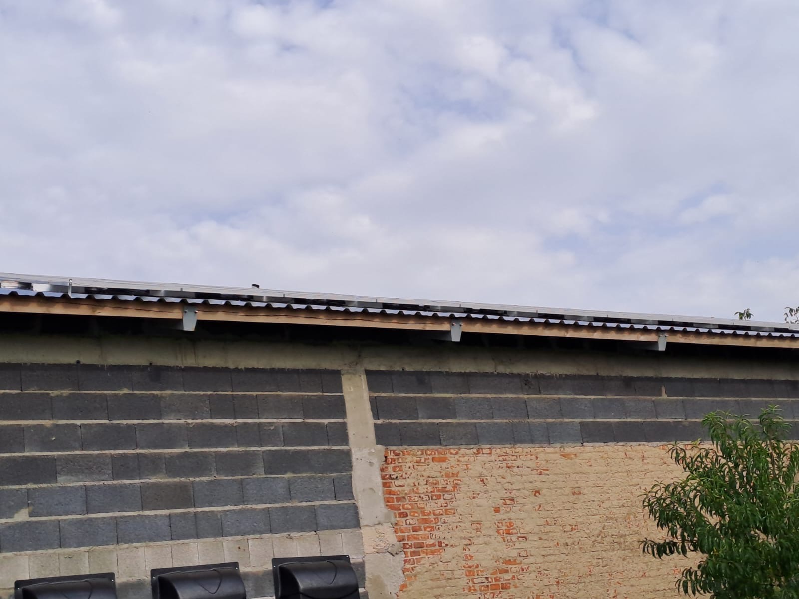 Montaż paneli słonecznych na dachu budynku gospodarczego z bloków betonowych i cegły, widoczne klamry mocujące konstrukcję.