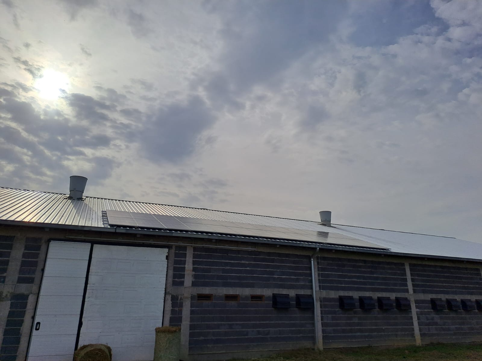 Panele słoneczne na dachu budynku gospodarczego z szarej cegły, widoczne kominy wentylacyjne i białe drzwi przesuwne.