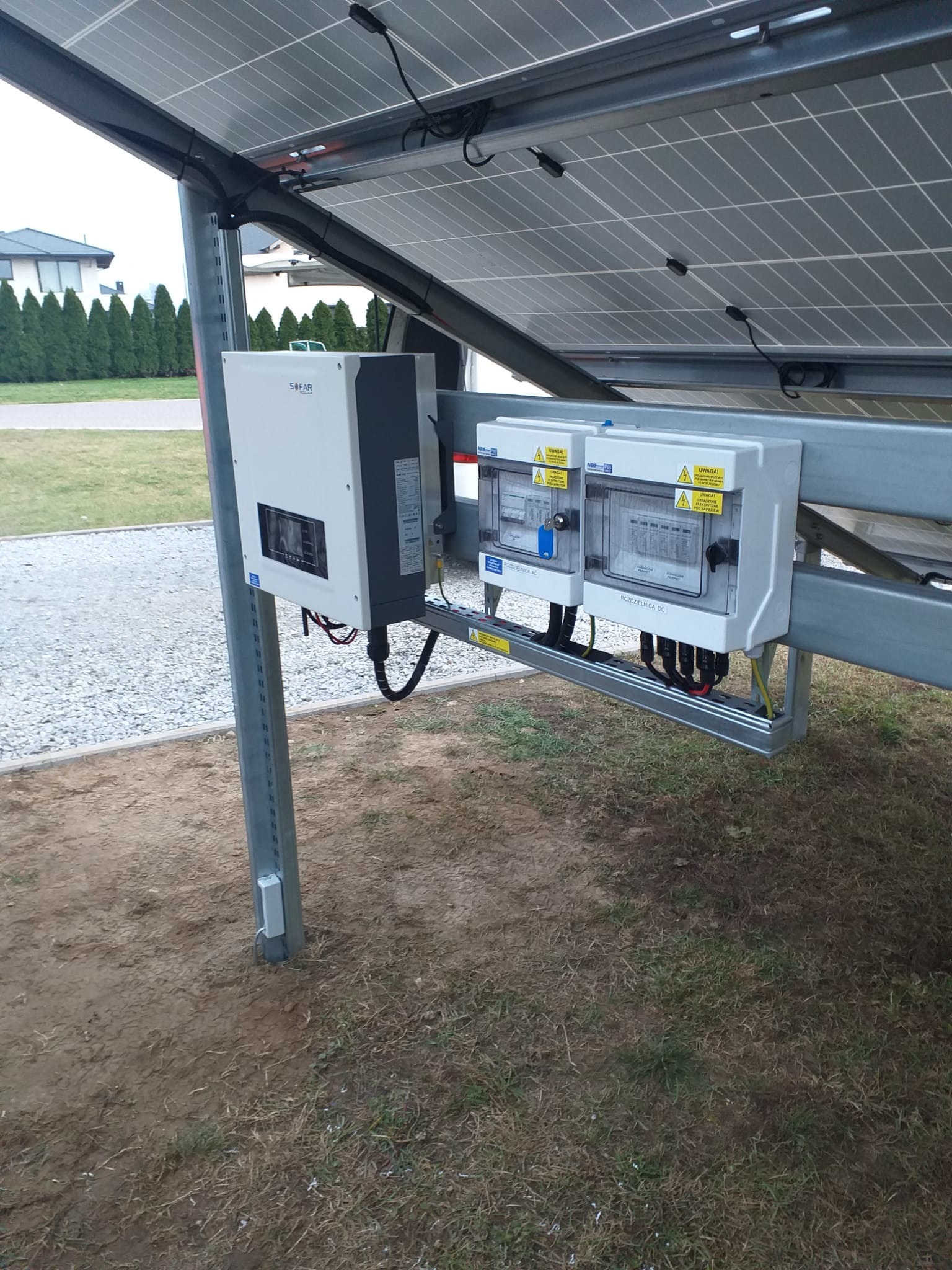 Instalacja paneli słonecznych z widocznym inwerterem SOFAR i rozdzielnicami DC/AC, zamontowana na metalowej konstrukcji w otoczeniu trawnika i budynku mieszkalnego.